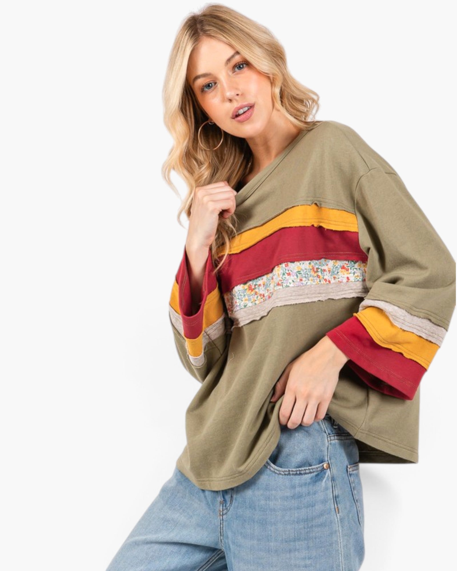 A woman modeling a loose olive green V-neck top with horizontal stripes in mustard yellow, red, and a multicolor floral lace panel, paired with light blue jeans against a white studio background.