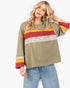 A woman modeling a loose olive green long-sleeve top with horizontal stripes in red, yellow, and a multicolor floral lace panel across the chest, paired with light blue jeans against a white studio background.