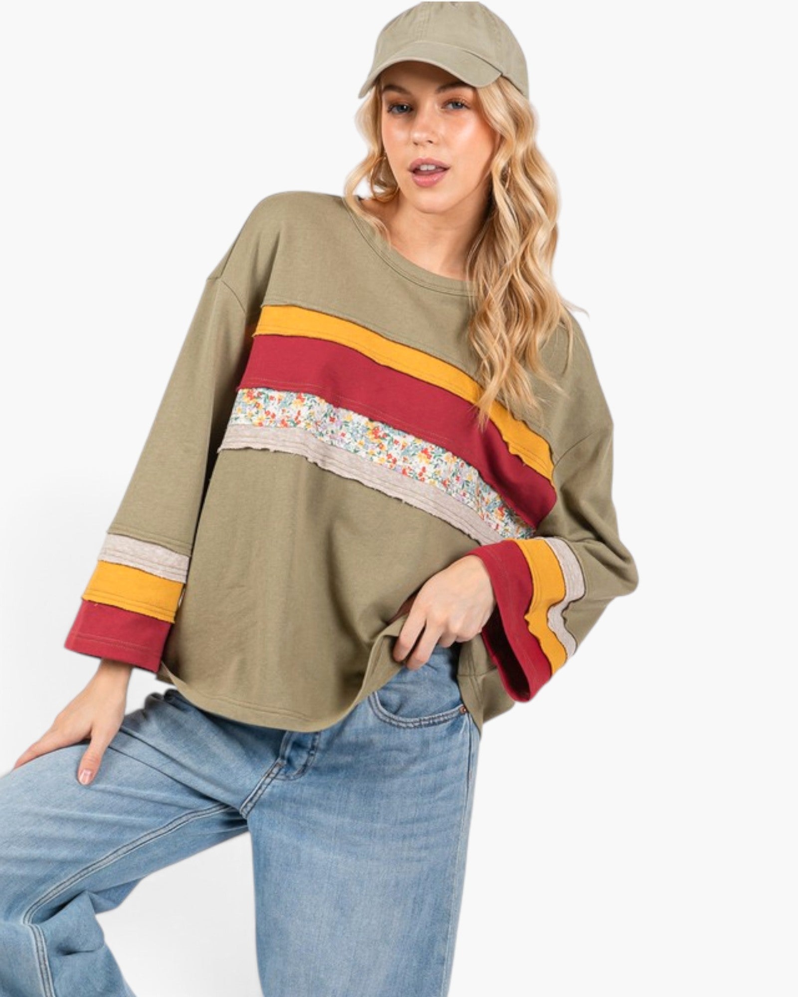 A woman wearing a loose olive green long-sleeve top with colorful horizontal stripes in yellow, red, and a floral pattern, paired with blue jeans and a matching olive green cap, against a plain white background.