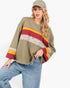 A woman wearing a loose olive green long-sleeve top with colorful horizontal stripes in yellow, red, and a floral pattern, paired with blue jeans and a matching olive green cap, against a plain white background.