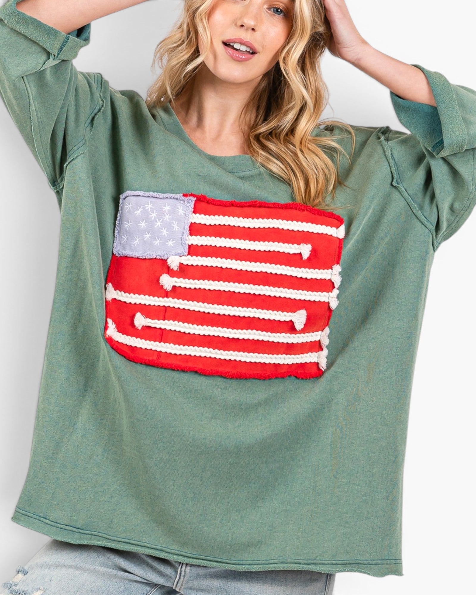 A woman wearing a sage green long-sleeve t-shirt with a large 3D knitted American flag patch on the front, paired with light-wash jeans against a plain white background.