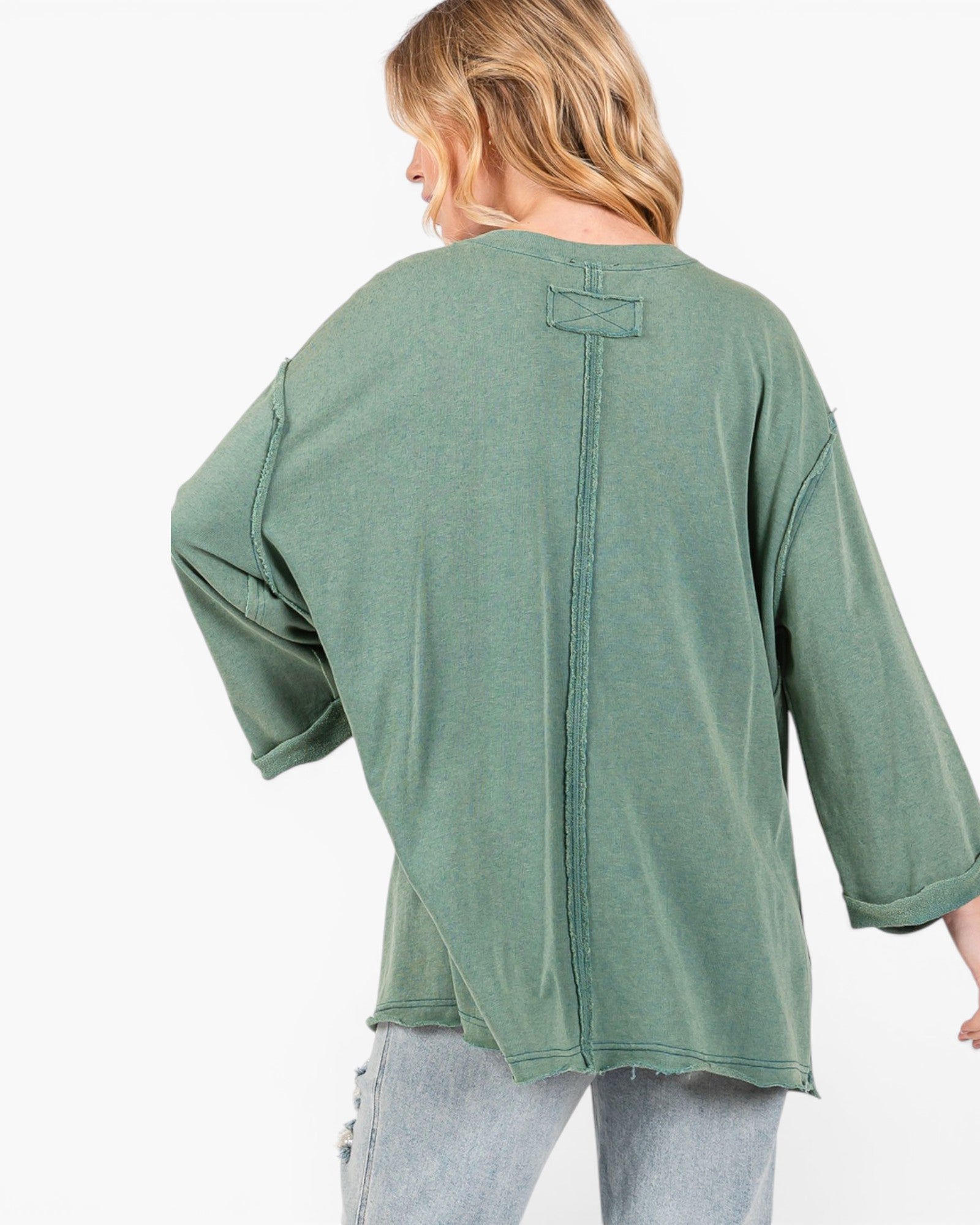 A woman shown from the back wearing an oversized sage green top with a vertical seam and tie detail at the neckline, paired with light wash jeans against a plain white background.