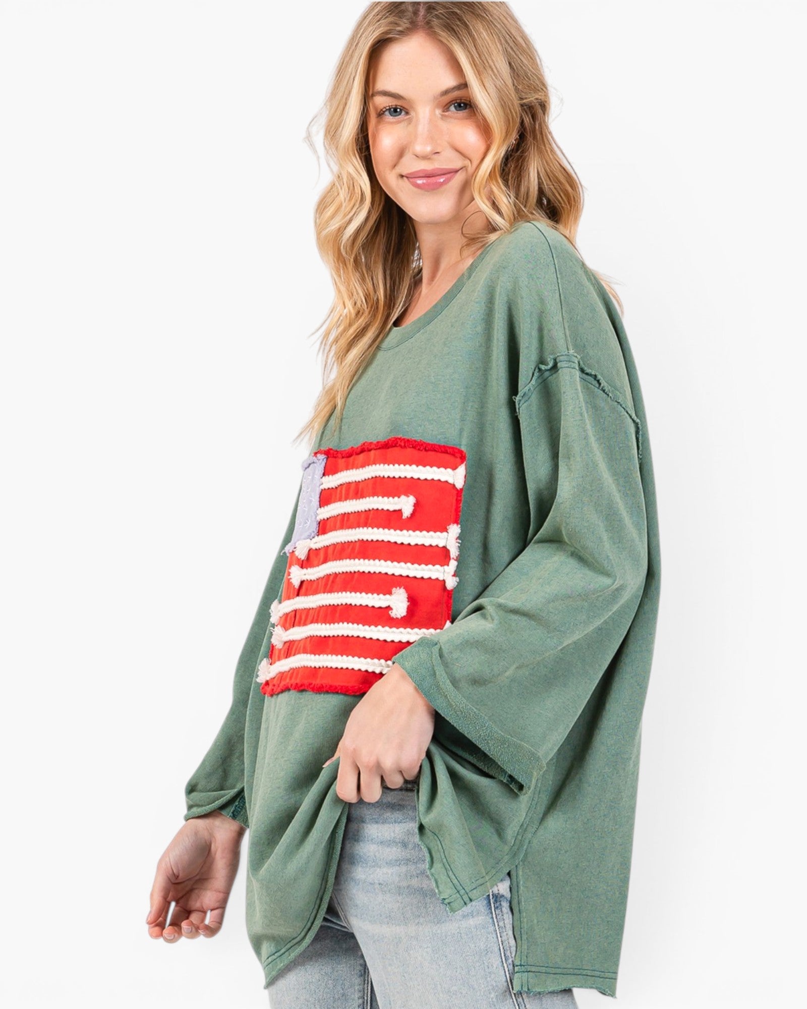 A woman wearing a sage green knit sweater with a red and white striped knit patch on the front, paired with light wash jeans, against a plain white background.