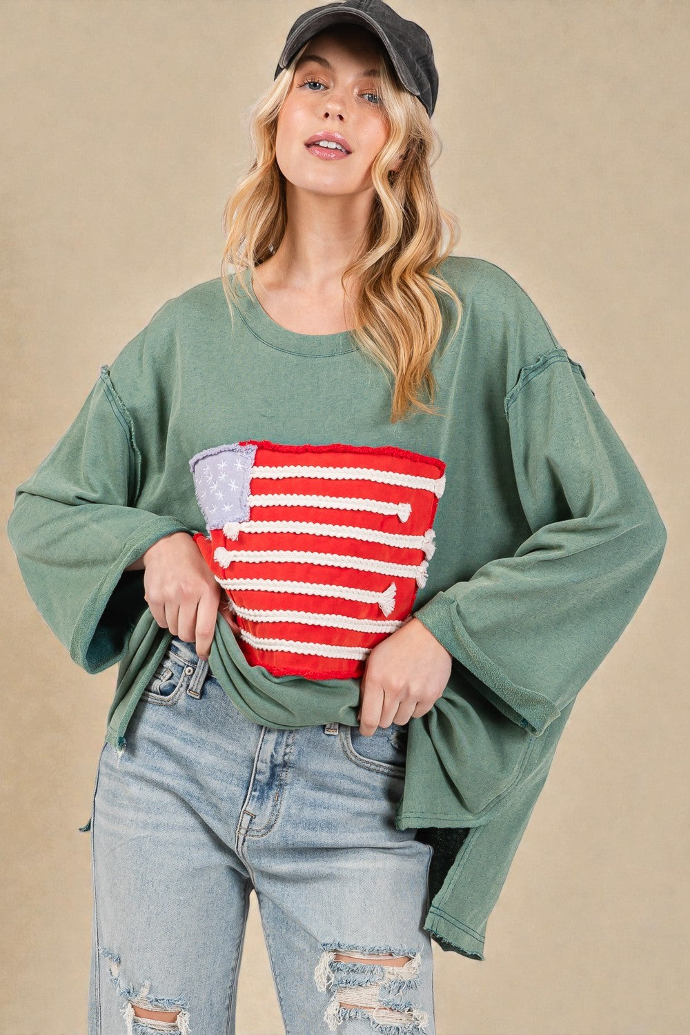 Woman wearing a green oversized sweater with an American flag design, blue jeans, and a cap on a beige background.