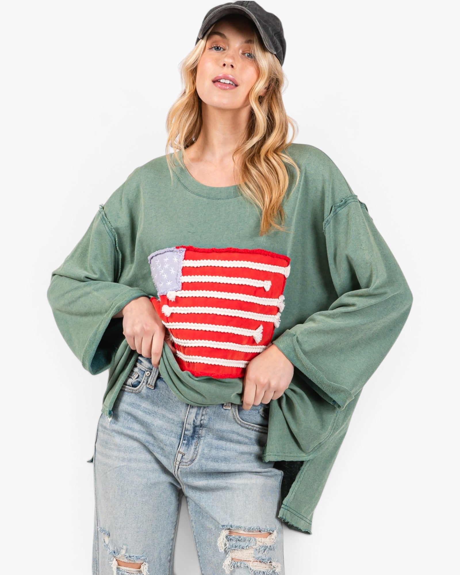 A woman wearing a green long-sleeve top with a red and white striped graphic on the front, paired with light-wash distressed jeans and a black baseball cap, against a plain white background.