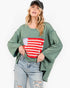 A woman wearing a green long-sleeve top with a red and white striped graphic on the front, paired with light-wash distressed jeans and a black baseball cap, against a plain white background.
