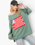 A woman modeling an oversized sage green knit top with a distressed American flag patch on the front, paired with ripped light-wash jeans and a black baseball cap, against a plain white background.