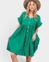 A woman wearing a bright green short-sleeve button-front dress with a gathered waist and a brown wide-brim hat, photographed against a light gray studio background.