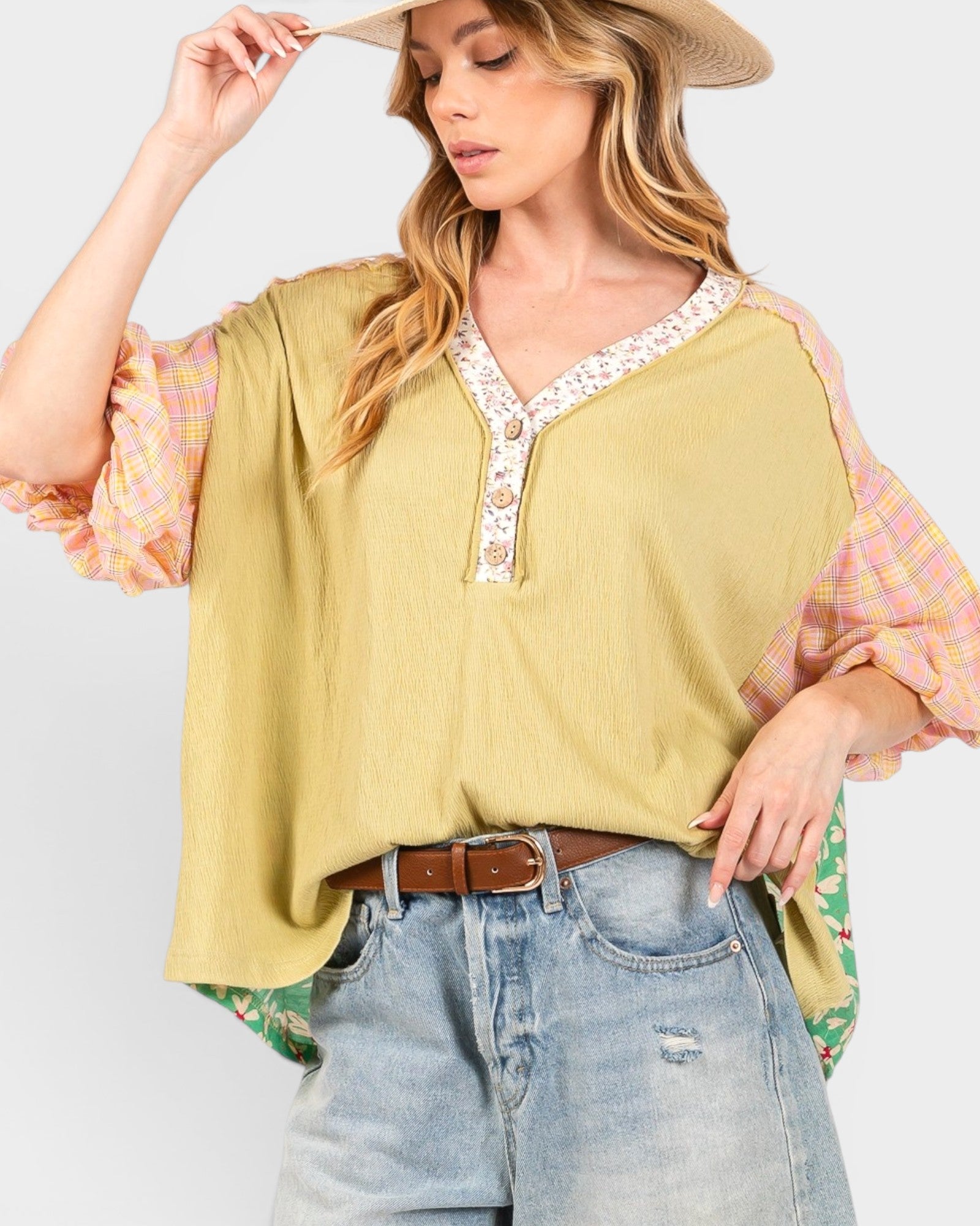 A woman wearing a loose olive yellow top with floral lace trim on the V-neck and ruffled pink plaid sleeves, styled with light-wash jeans, a brown belt, and a straw hat against a light gray background.