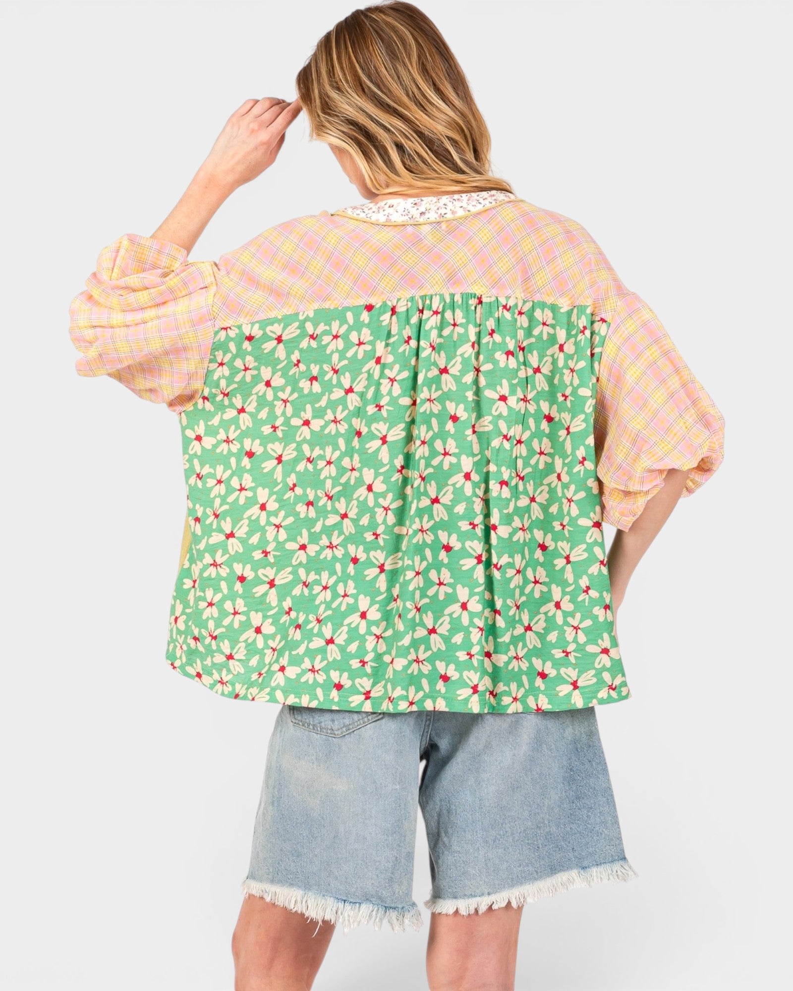 A woman shown from the back wearing a patchwork blouse with a green floral print lower section, a pink plaid upper section, and light blue denim shorts against a light gray background.