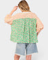 A woman shown from the back wearing a patchwork blouse with a green floral print lower section, a pink plaid upper section, and light blue denim shorts against a light gray background.
