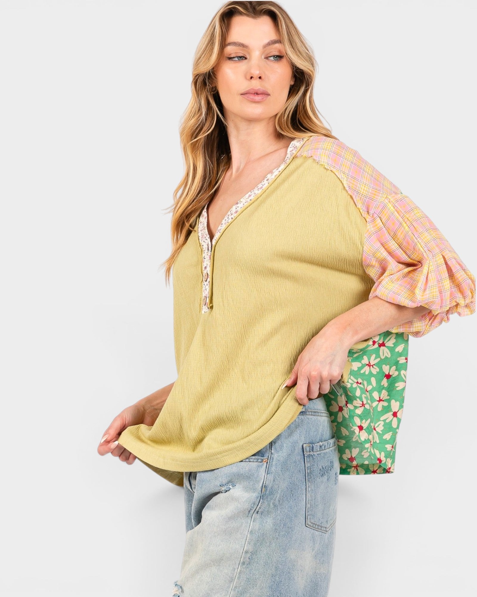 A woman modeling a yellow top with pink plaid puff sleeves and a floral green bag, set against a plain light gray background.