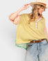 A woman wearing a yellow-green sleeveless top with a floral-patterned undershirt, light-wash jeans, a brown belt, and a straw hat, photographed against a light gray studio background.