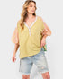 A woman modeling a yellow-green short-sleeve blouse with pink plaid puff sleeves and a white lace-trimmed V-neckline, paired with light-wash denim shorts against a light gray studio background.