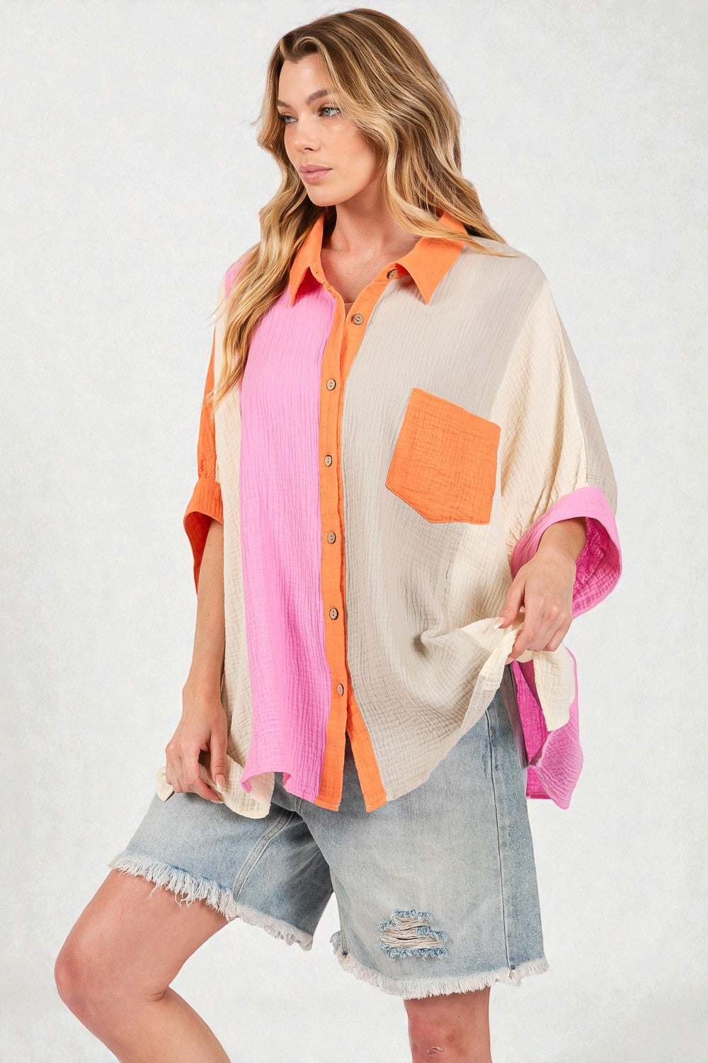 Woman wearing a colorful layered outfit with a pink shirt, beige cardigan, and denim shorts on a white background.