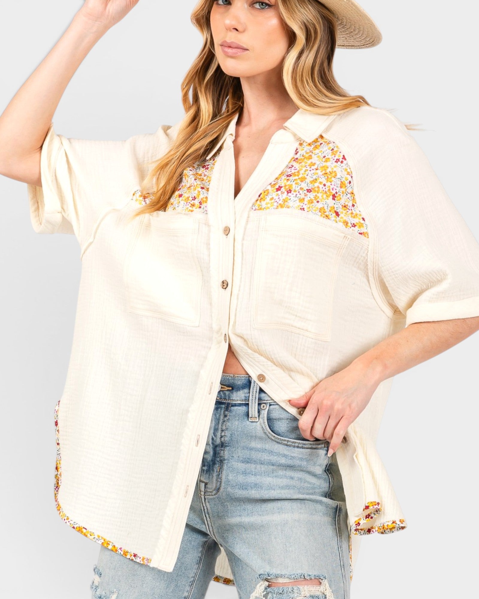 A woman wearing a cream-colored short-sleeve button-up shirt with yellow, orange, and white floral patchwork details on the chest and hem, paired with light-wash distressed jeans and a straw hat.