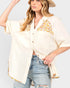 A woman wearing a cream-colored short-sleeve button-up shirt with yellow, orange, and white floral patchwork details on the chest and hem, paired with light-wash distressed jeans and a straw hat.
