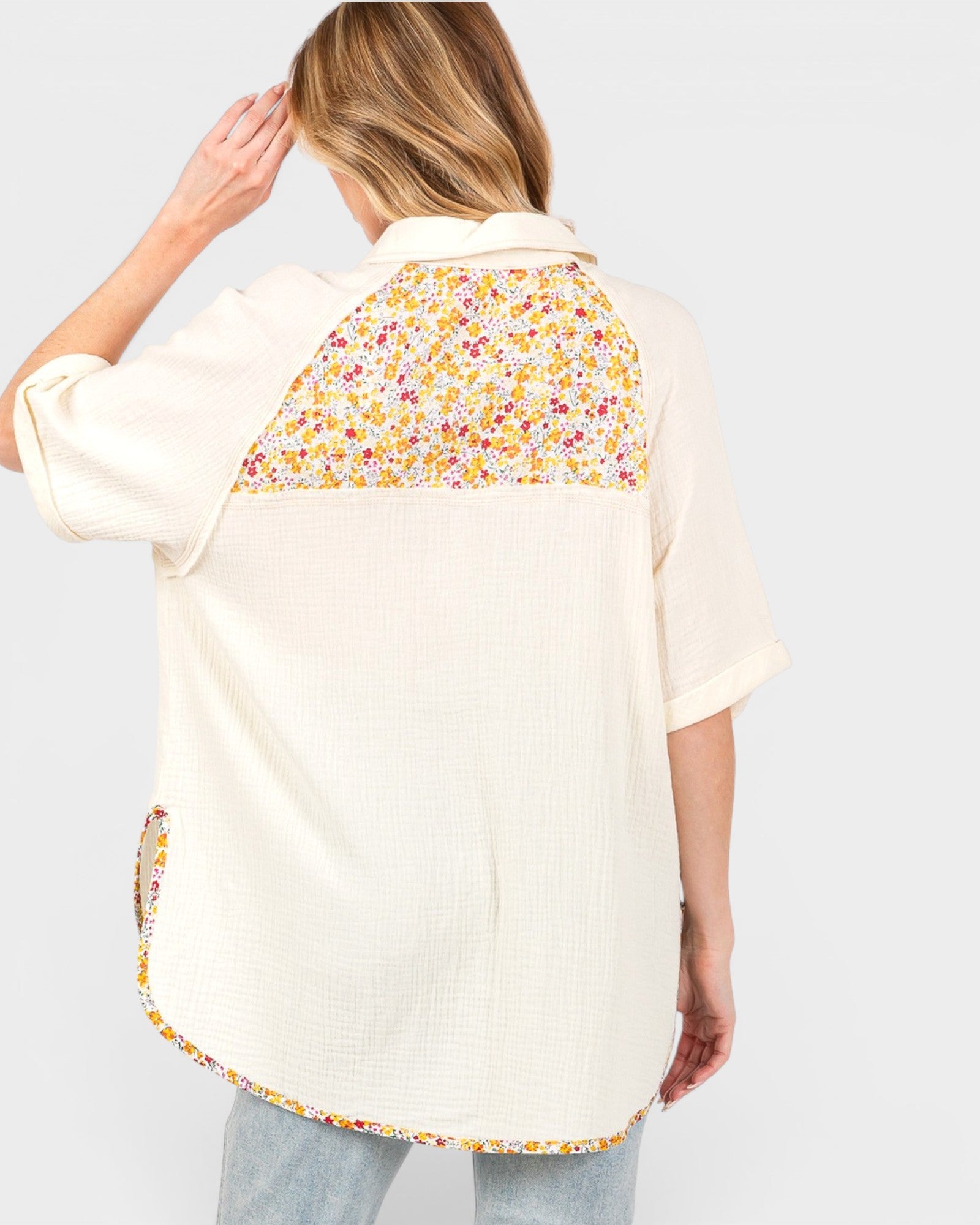 A woman shown from the back wearing a cream-colored short-sleeve shirt with a colorful floral pattern panel across the upper back and matching floral trim along the hem, paired with light blue jeans against a light gray background.