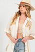 Woman wearing a cream-colored embroidered shirt, denim shorts, and a straw hat on a white background