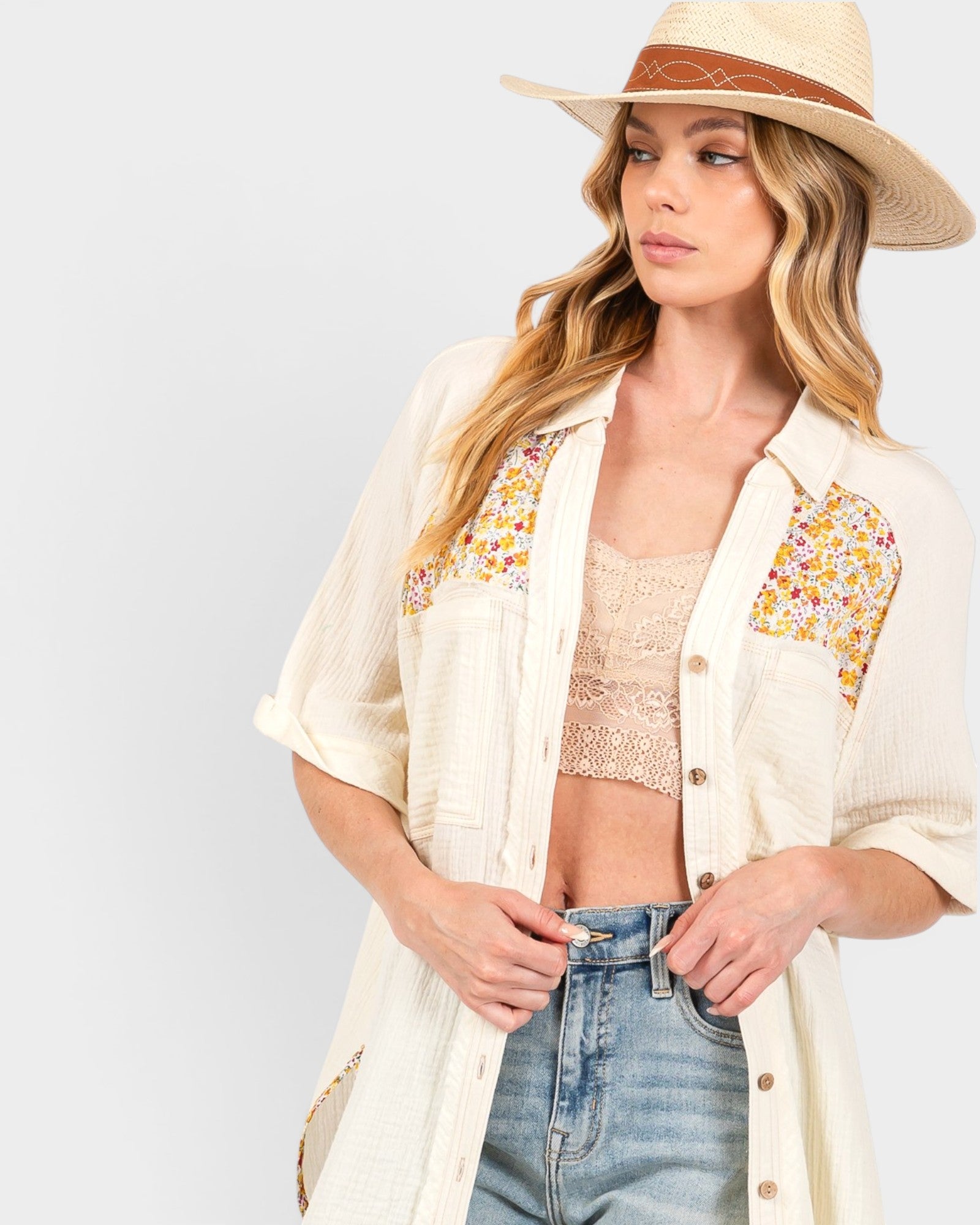 A woman wearing a cream-colored, open-front button-up shirt with colorful floral embroidery on the chest pockets, styled with a straw hat, beige lace crop top, and blue jeans against a light gray background.