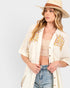 A woman wearing a cream-colored, open-front button-up shirt with colorful floral embroidery on the chest pockets, styled with a straw hat, beige lace crop top, and blue jeans against a light gray background.