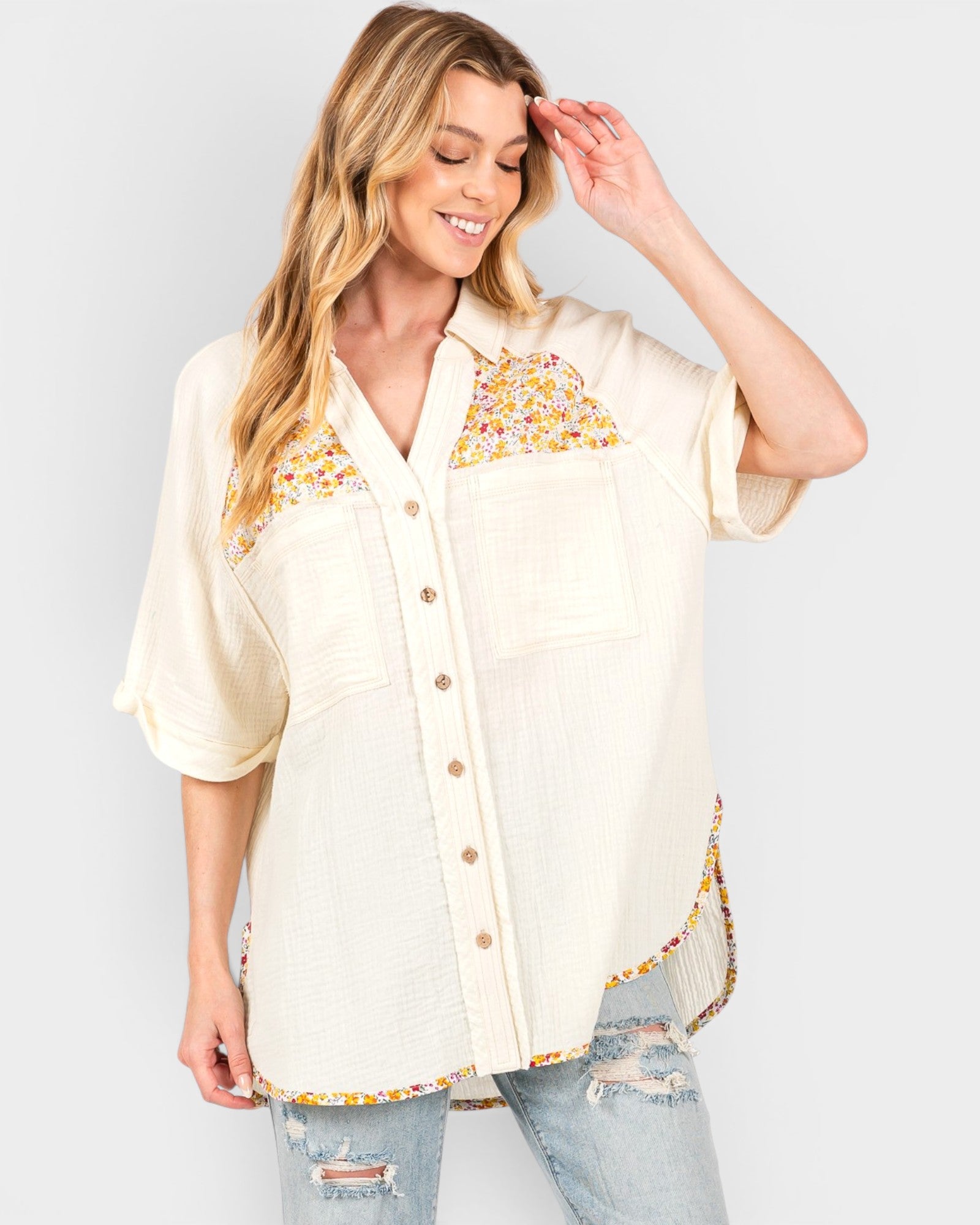 A woman wearing a cream-colored short-sleeve button-up shirt with yellow and orange floral embroidery on the chest and a matching embroidered hem, paired with distressed light-wash jeans, against a light gray studio background.