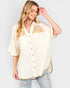 A woman wearing a cream-colored short-sleeve button-up shirt with yellow and orange floral embroidery on the chest and a matching embroidered hem, paired with distressed light-wash jeans, against a light gray studio background.