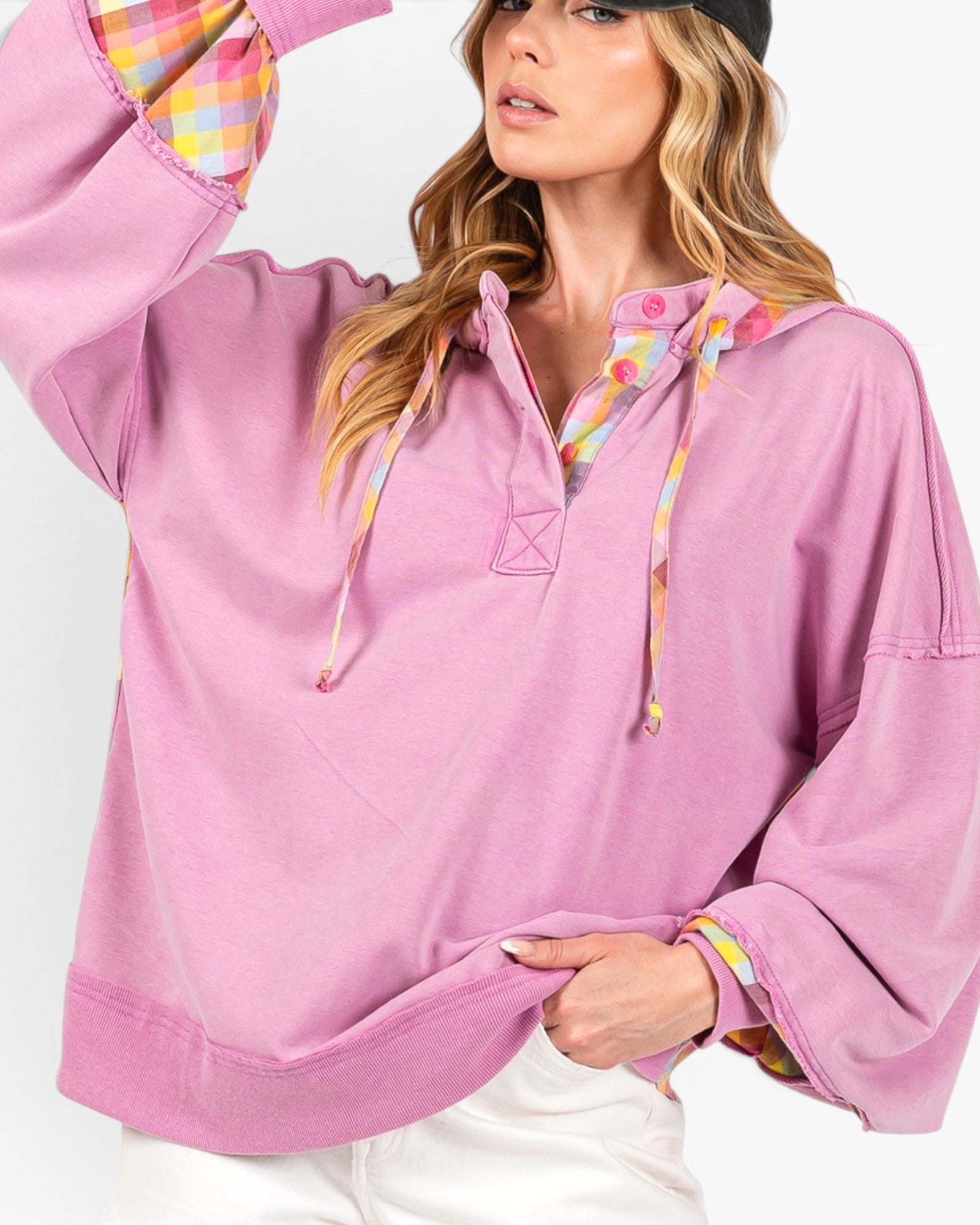 A woman wearing an oversized light pink pullover hoodie with colorful patterned drawstrings and cuffs, paired with white pants against a plain light background.