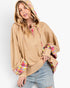 A woman wearing a tan oversized pullover hoodie with colorful plaid lining details on the hood, cuffs, and inner placket, paired with light-wash distressed jeans against a plain white studio background.