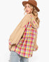 A woman wearing a beige oversized hoodie with a colorful pink, yellow, and blue plaid shirt layered underneath, paired with a brown wide-brim hat and light-wash jeans, photographed against a plain white background.