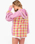 A woman wearing an oversized hoodie with a pink base and a yellow, pink, and white plaid pattern on the back and sleeves, shown from a side-back angle against a plain white background.