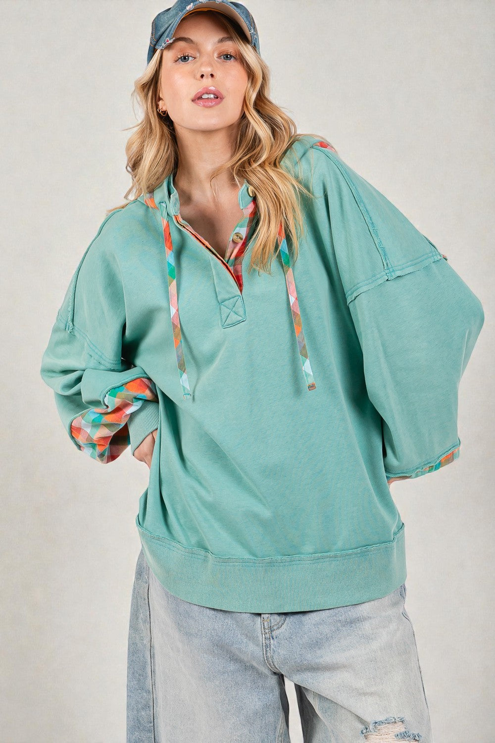 Woman wearing a teal hoodie with colorful accents and a blue cap against a plain background