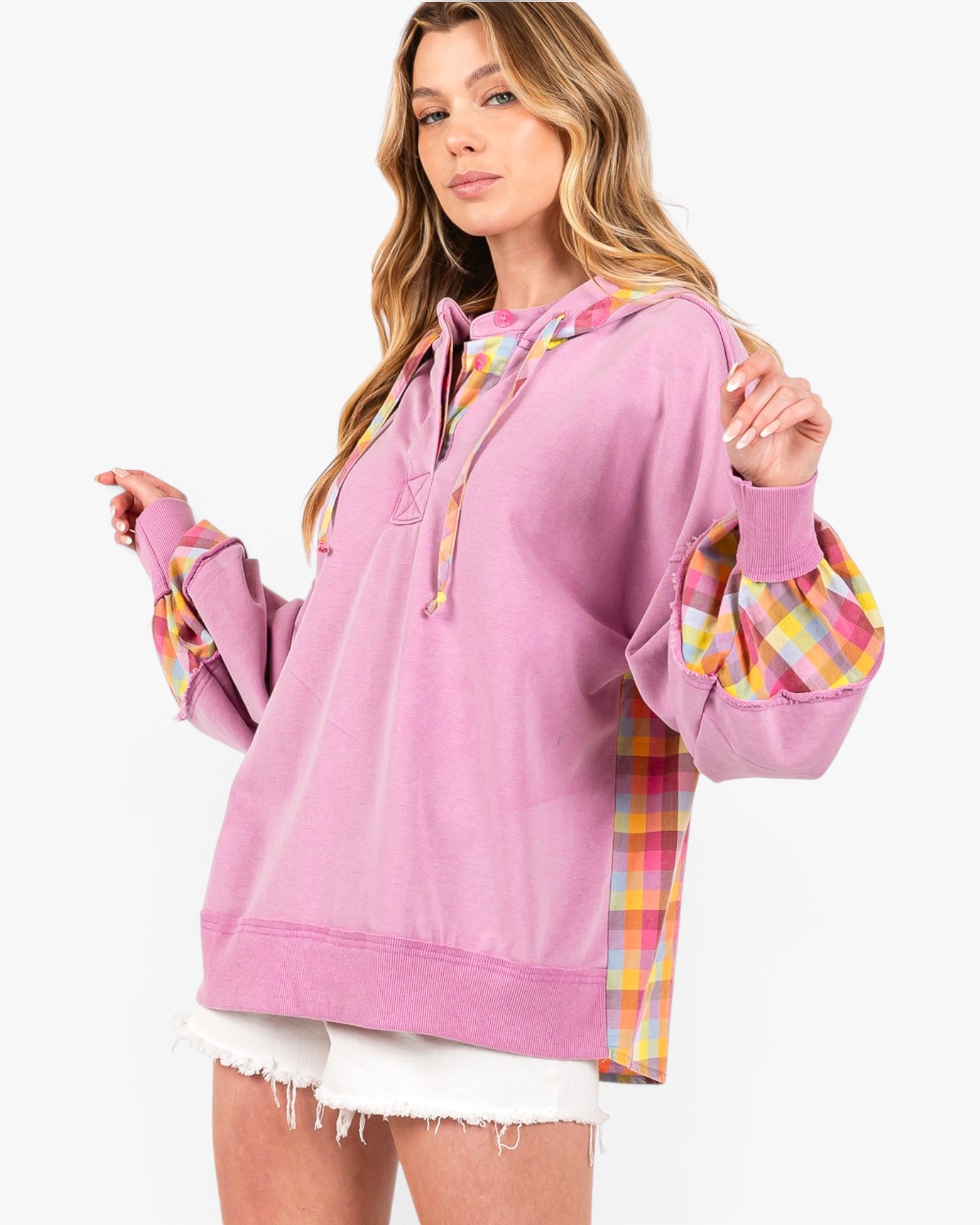 A woman wearing a lavender-purple pullover hoodie with colorful plaid trim details on the hood, cuffs, and side panels, styled with white frayed denim shorts against a plain white background.