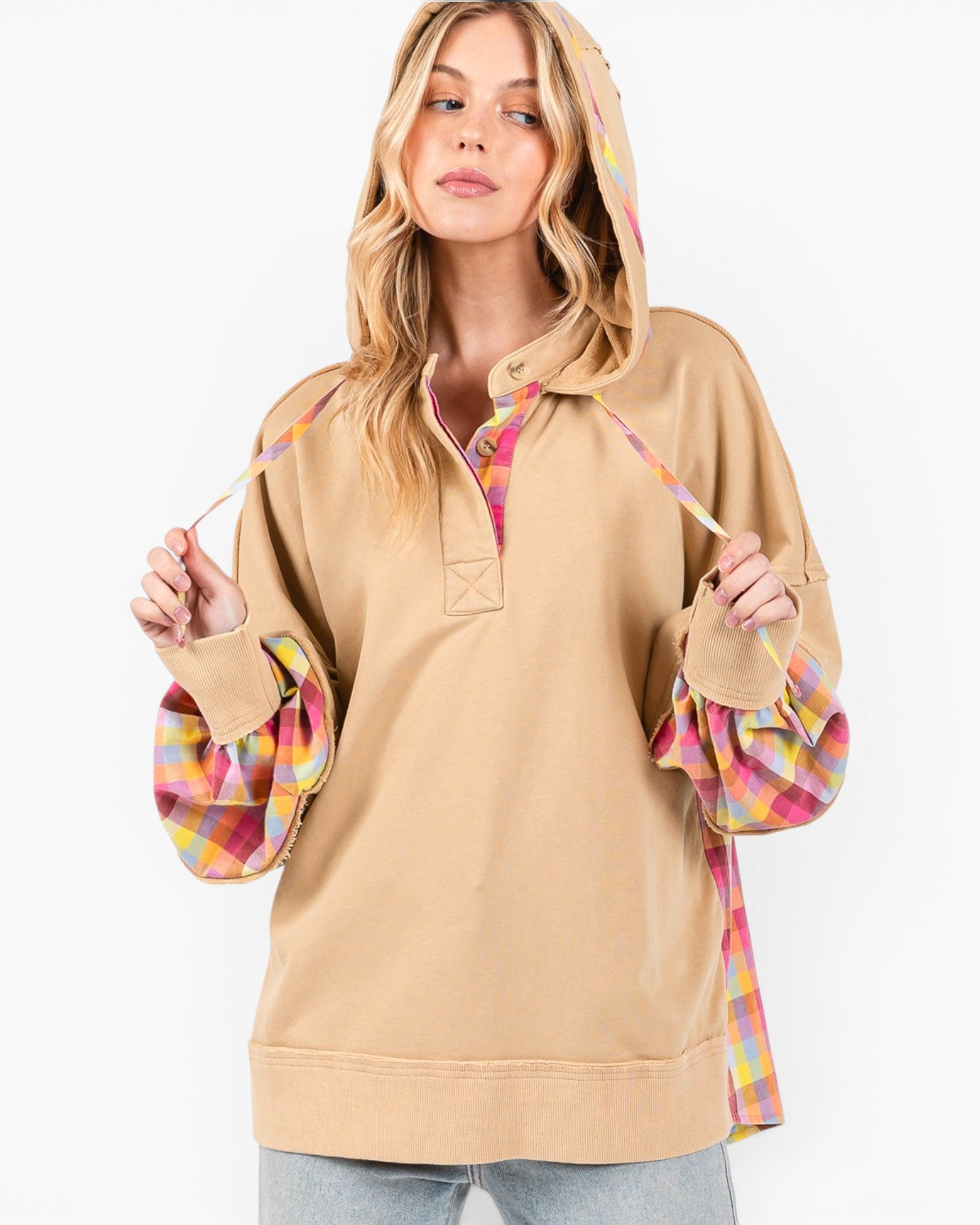 A woman wearing a beige pullover hoodie with colorful plaid trim and ruffled sleeves, shown against a plain light gray studio background.
