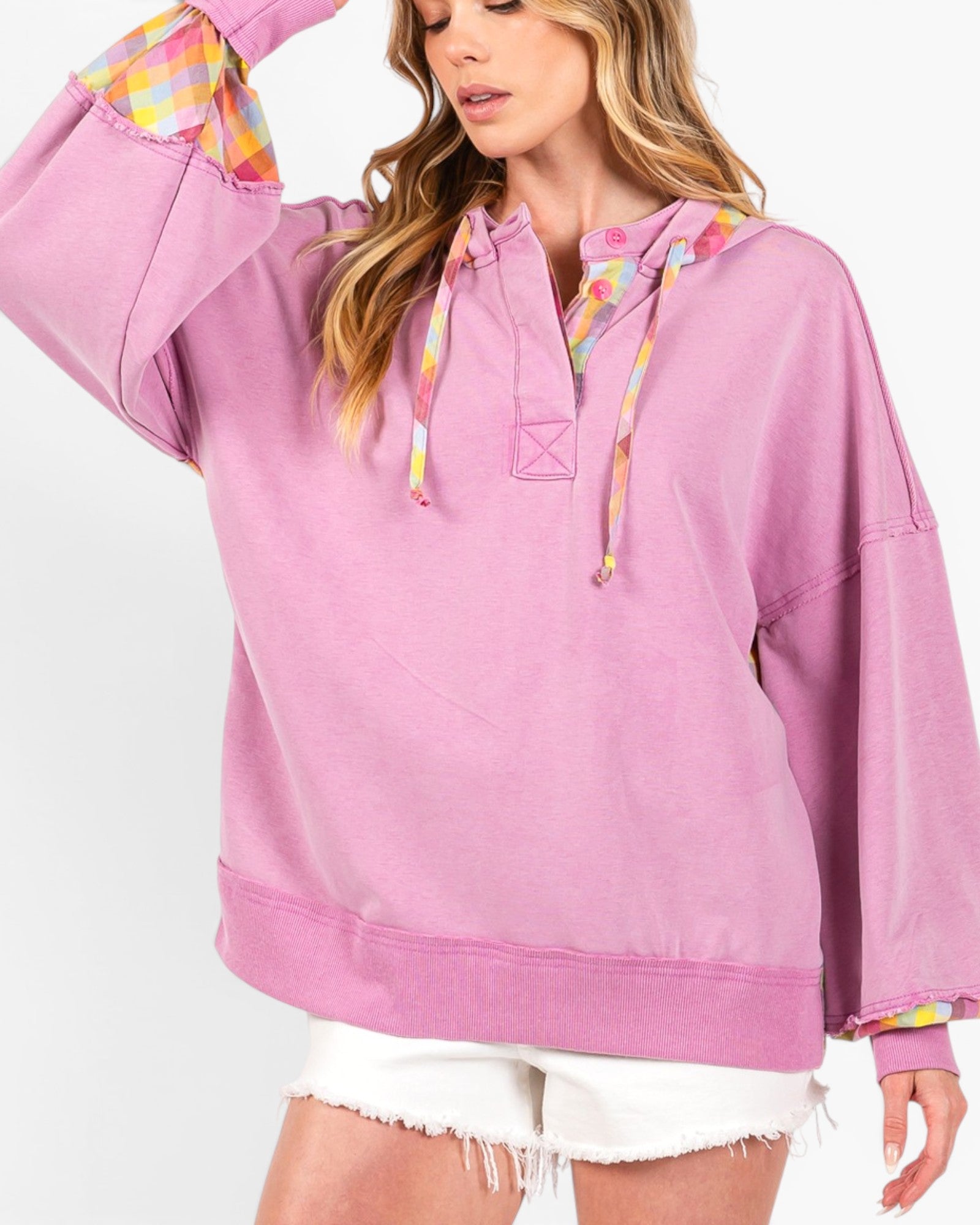 A woman wearing a pink oversized hoodie with a colorful plaid pattern lining the hood and cuffs, paired with white frayed denim shorts against a light gray studio background.