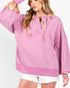 A woman wearing a pink oversized hoodie with a colorful plaid pattern lining the hood and cuffs, paired with white frayed denim shorts against a light gray studio background.