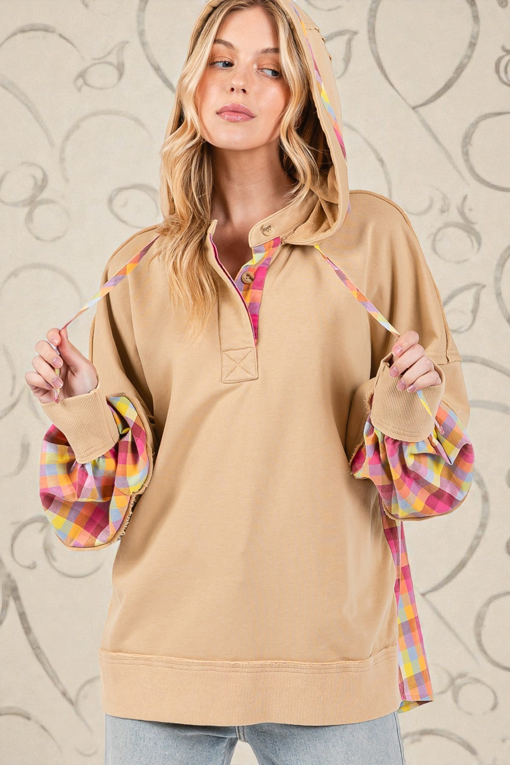 Woman wearing a beige pullover with plaid sleeves against a decorative wall.