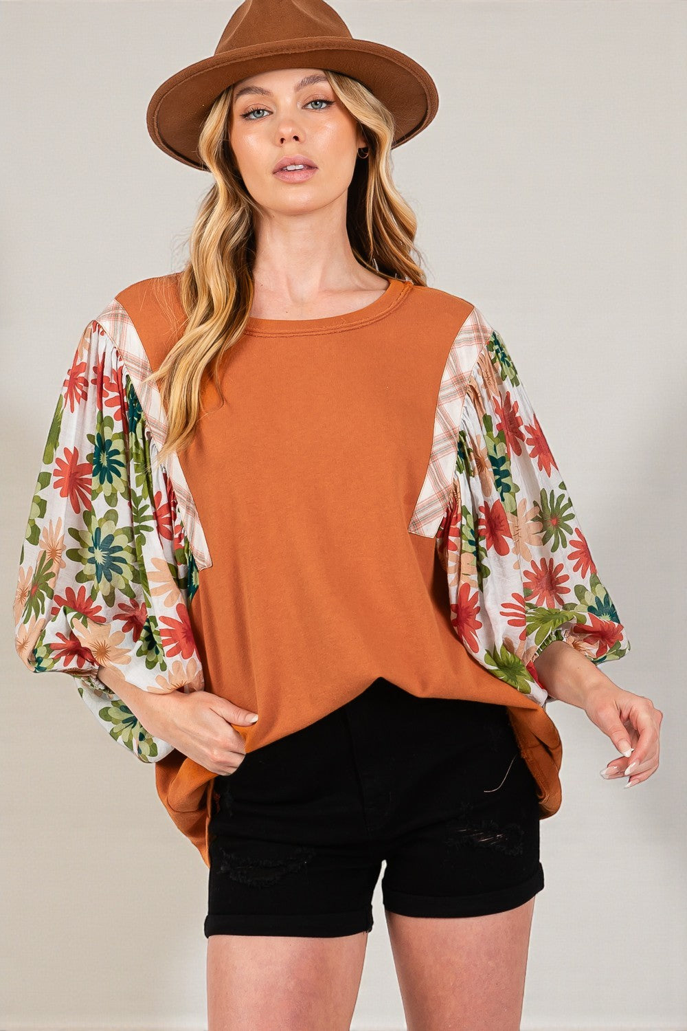 Woman wearing a rust-colored top with floral sleeves, black shorts, and a brown hat on a plain background