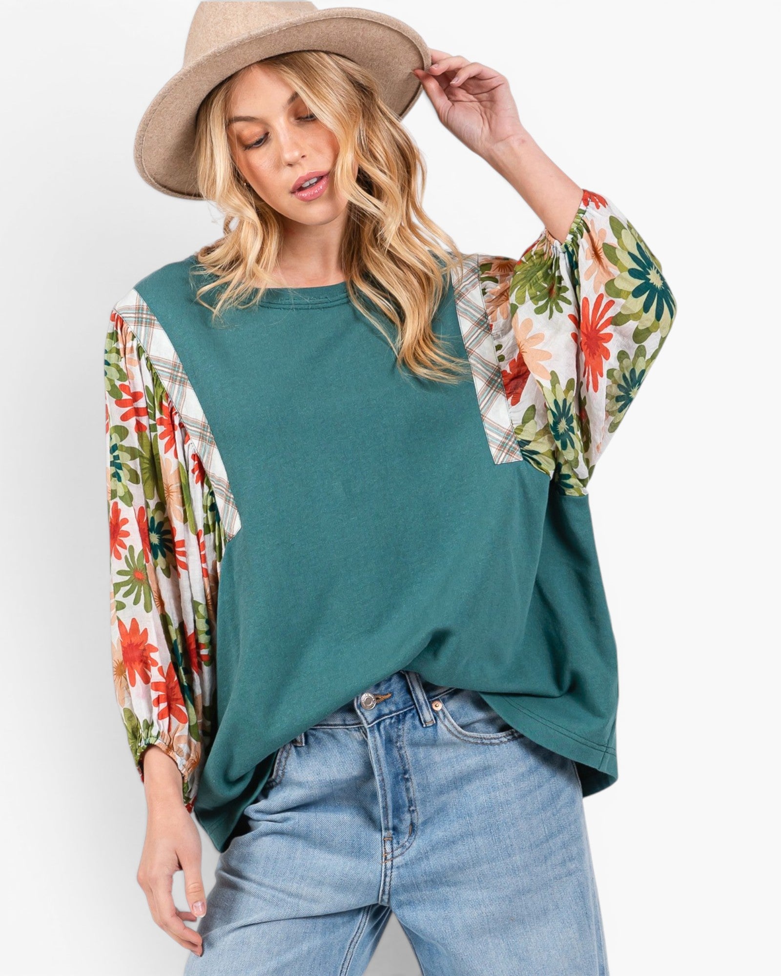 A woman wearing a teal green top with floral-patterned bell sleeves, paired with light blue jeans and a beige wide-brim hat, against a plain white background.