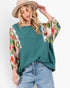 A woman wearing a teal green top with floral-patterned bell sleeves, paired with light blue jeans and a beige wide-brim hat, against a plain white background.
