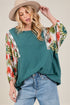 Woman wearing a teal top with floral sleeves and light blue jeans, holding a beige hat.