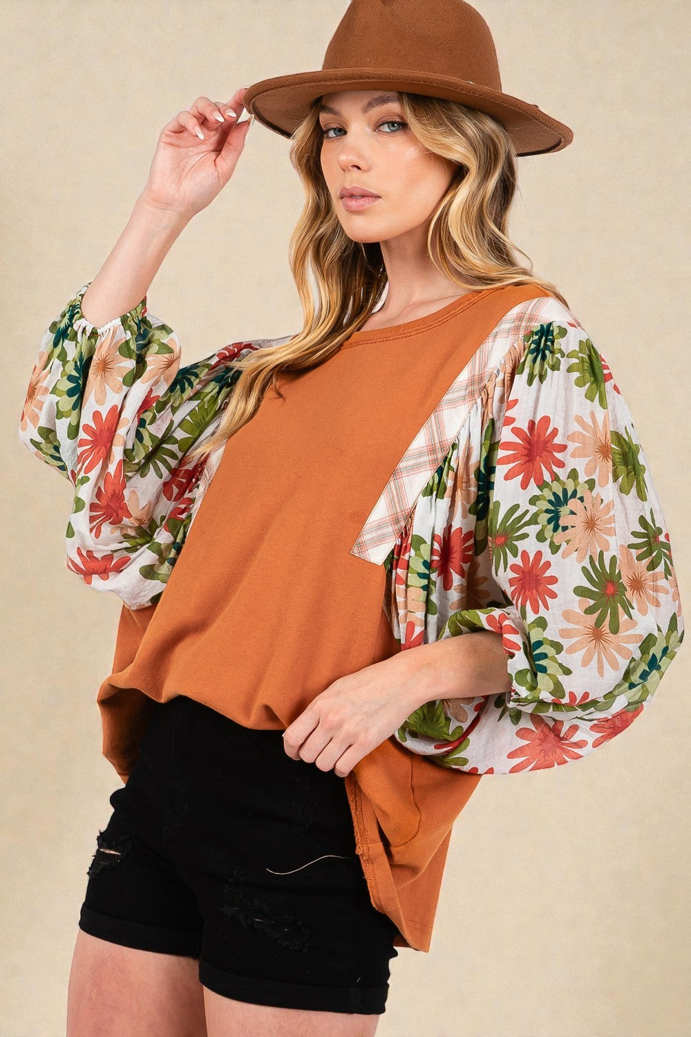 Woman wearing a floral kimono over a rust-colored top and black shorts, holding a brown hat.