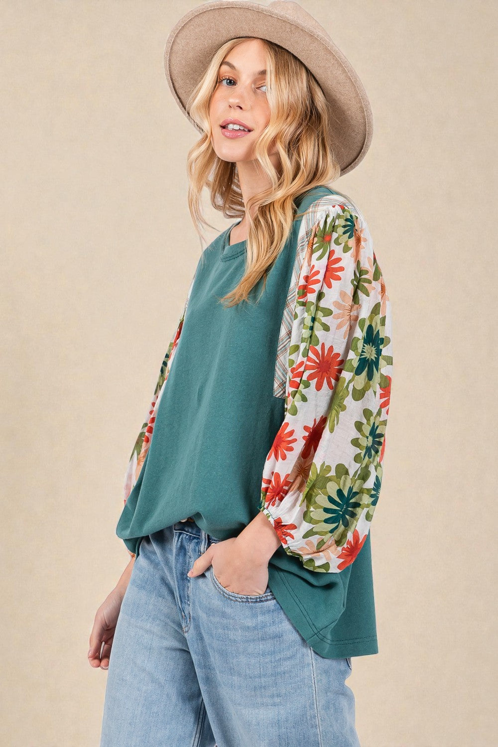 Woman wearing a teal top with floral sleeves and a beige hat against a beige background