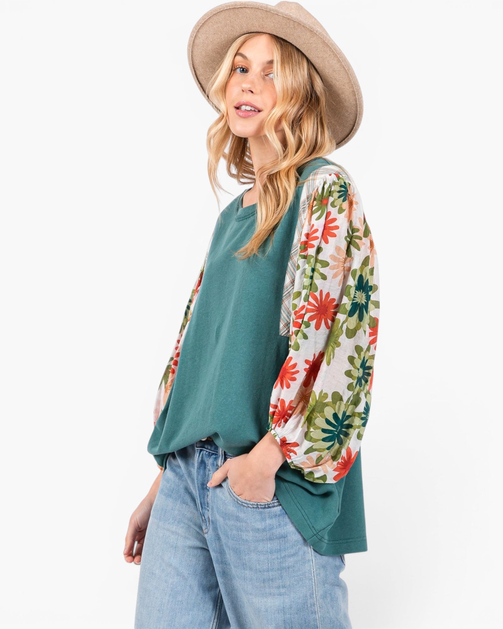 A woman wearing a teal green top with long white sleeves featuring a red, green, and orange floral print, paired with light blue jeans and a beige wide-brim hat, against a plain white background.