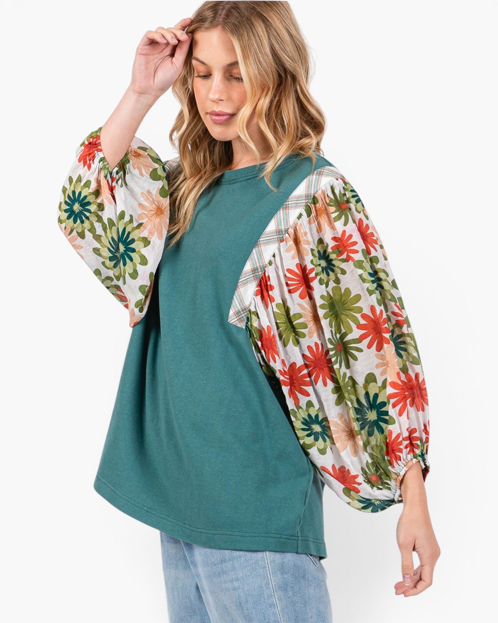 A woman wearing a teal raglan-style top with dramatic floral puff sleeves in red, green, and cream, paired with light blue jeans against a plain white background.