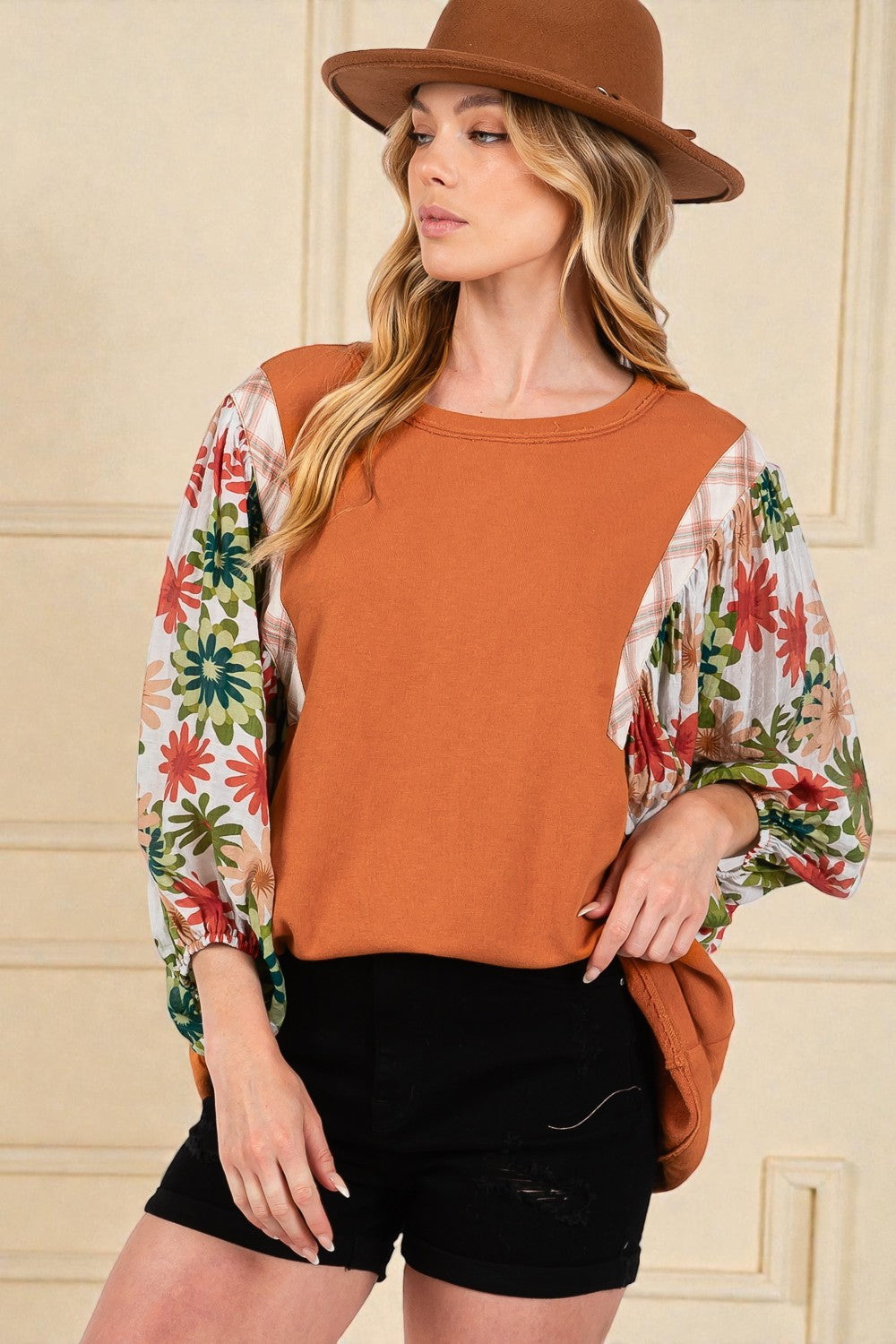 Woman wearing a rust-colored top with floral sleeves and a brown hat indoors.