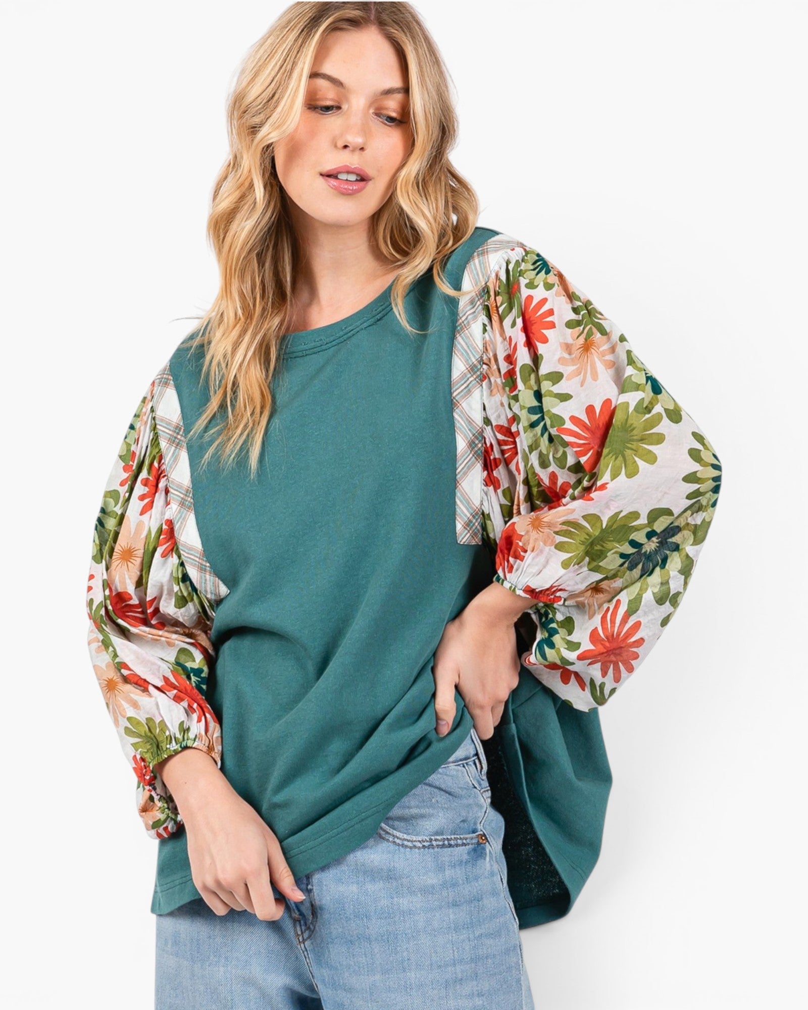 A woman wearing a teal green crewneck sweatshirt with dramatic, long balloon sleeves featuring a colorful floral and plaid print, paired with light blue jeans against a plain white background.