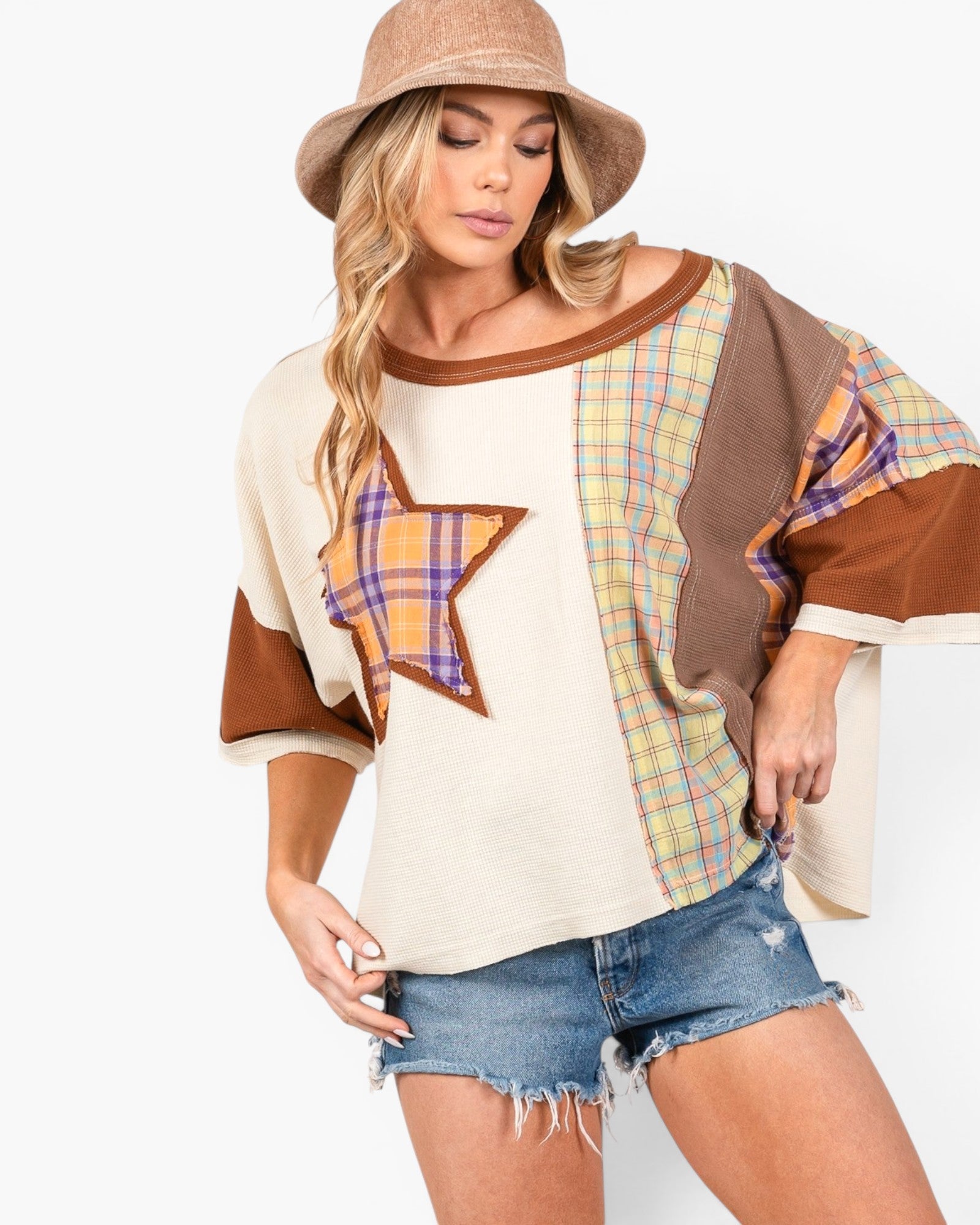 A woman modeling a bohemian-style patchwork top with a large star patch on the front, featuring cream, brown, and plaid patterns, paired with denim shorts and a tan hat against a white background.