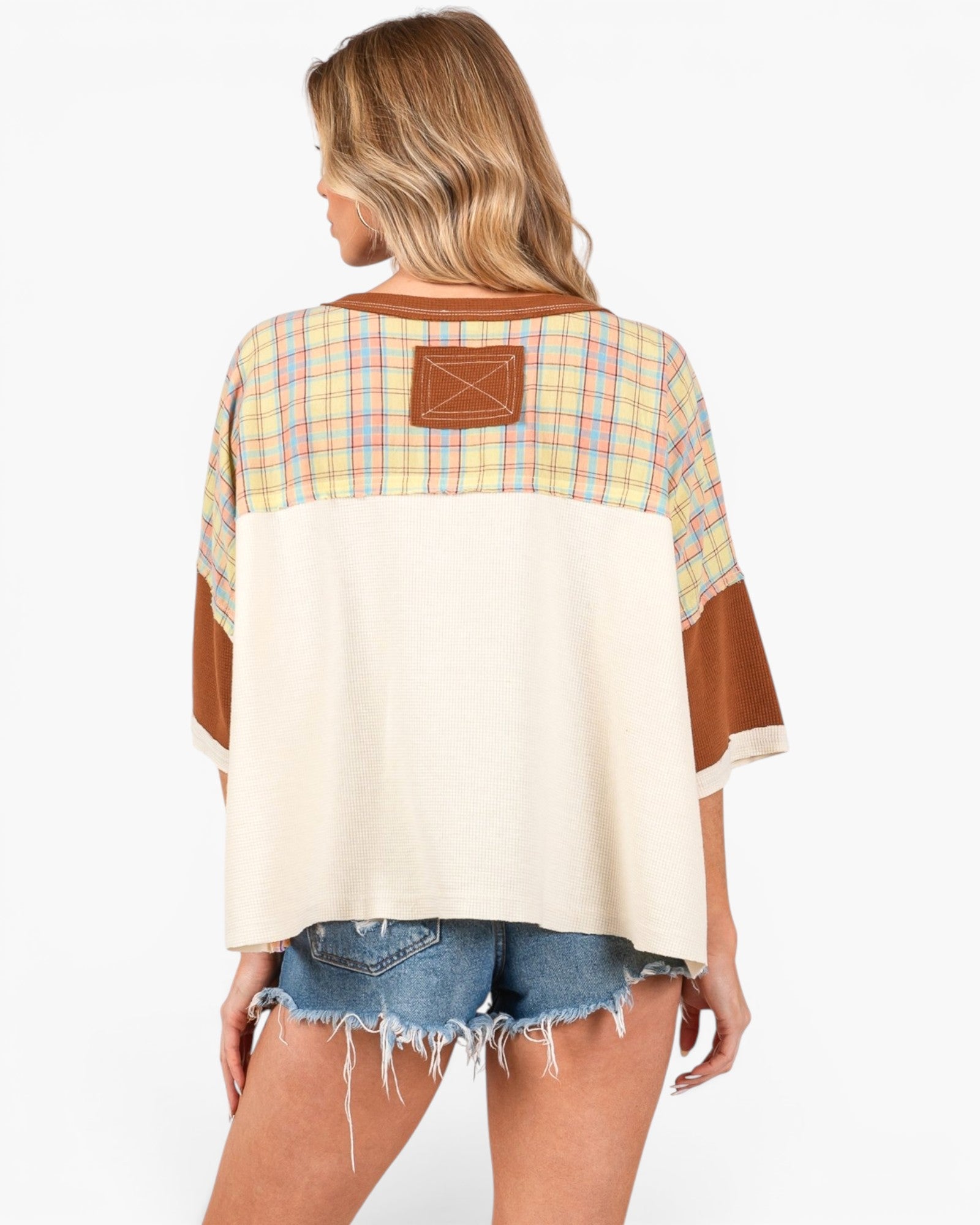 A woman shown from the back wearing a loose, color-block top with a cream body, a yellow and blue plaid yoke, and brown sleeve panels and a brown patch on the back, paired with denim shorts against a light gray studio background.