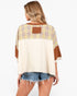 A woman shown from the back wearing a loose, color-block top with a cream body, a yellow and blue plaid yoke, and brown sleeve panels and a brown patch on the back, paired with denim shorts against a light gray studio background.